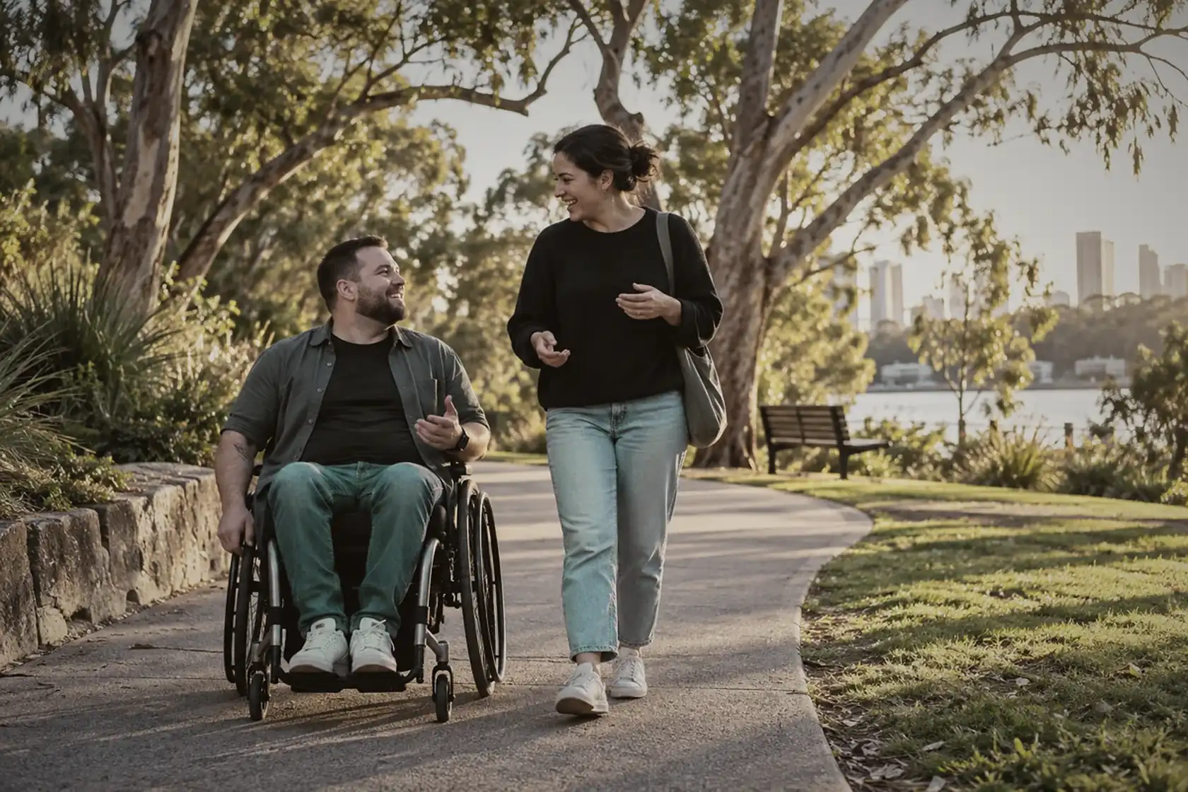 A man using a wheelchair and a friend chatting on a leafy park path at golden hour