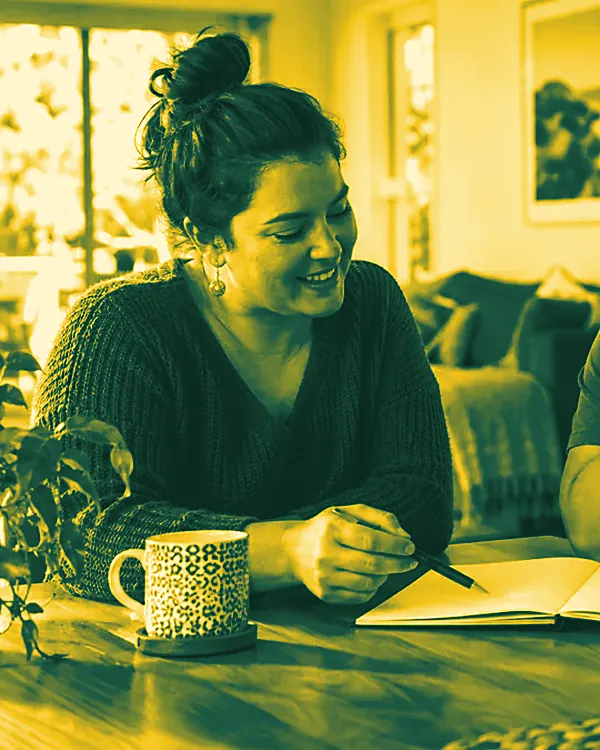 Illustrative photo: a smiling support worker writing in a notebook at a kitchen table